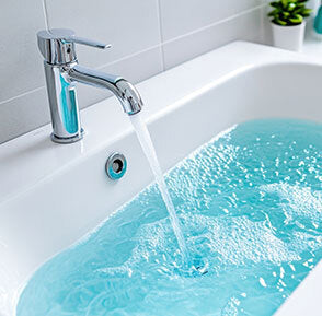 A chrome bathroom faucet running with clear water into a white sink filled with bright blue water, set against a light tiled wall.
