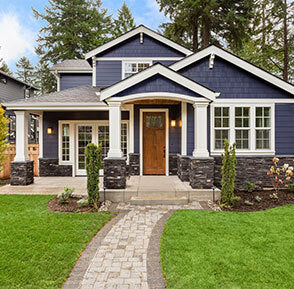 A two-story residential house exterior with dark blue siding, white trim, a wooden front door, stone accents, a brick walkway, and a green lawn with trees in the background.