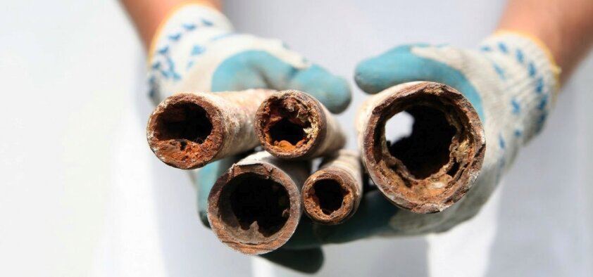 Gloved hands hold old, corroded water pipes with heavy limescale buildup.